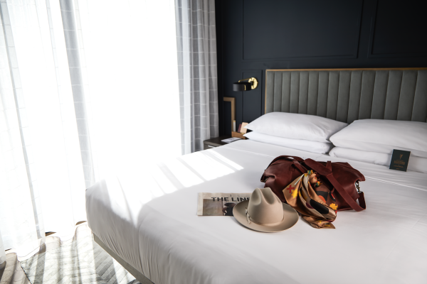 A hotel room with a neat bed, magazine, hat, bag, and scarf on it, beside a window with sheer curtains letting in natural light.