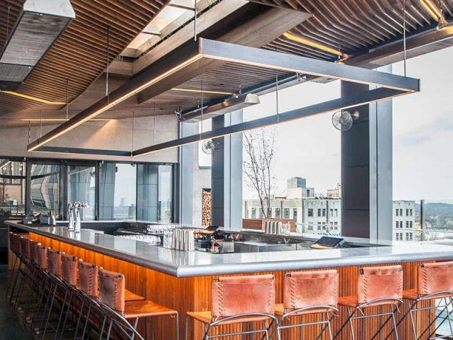 A bar with a long counter and stools, under a unique ceiling with hanging lights.