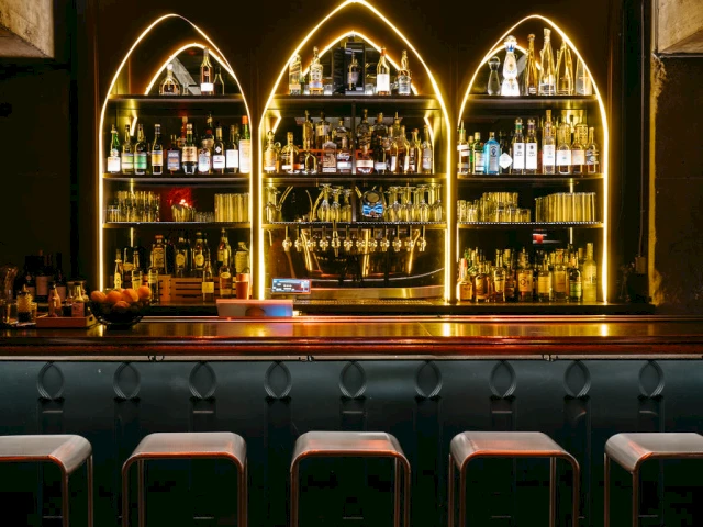 A dimly lit bar with a wooden counter, metal stools, and three arched shelving units filled with bottles and glasses, highlighted by neon lights.