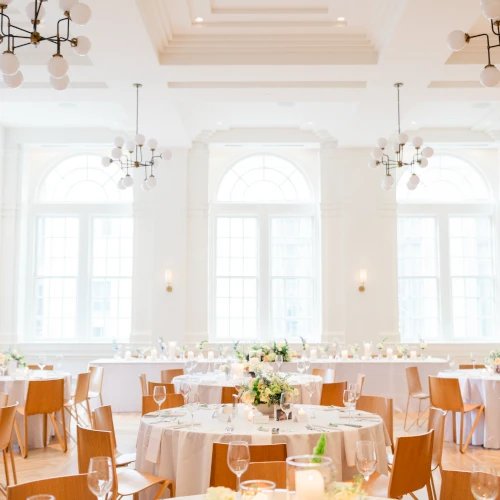 A brightly lit dining room with round tables set for a meal, featuring white tablecloths, wooden chairs, and elegant lighting fixtures.