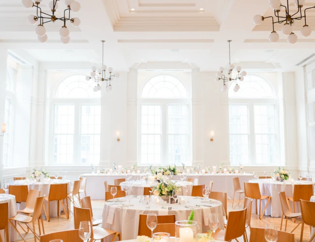 A brightly lit, elegantly set dining room with round tables, wood chairs, and floral arrangements.