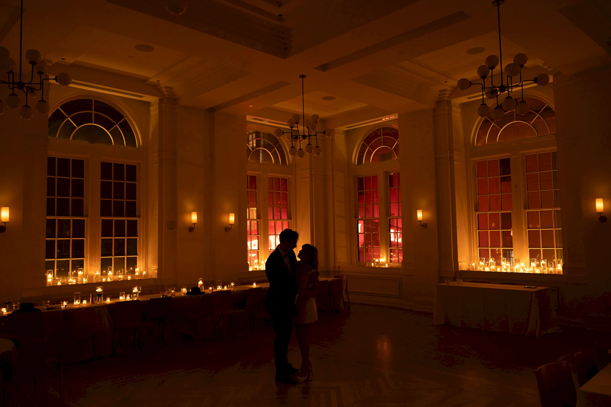 a dimly lit room with a couple in the center, surrounded by tables and chairs, with windows and chandeliers