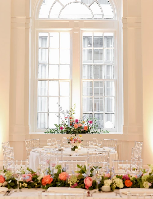 A formal dining room with a long table set for a meal, surrounded by clear chairs and decorated with flower arrangements under tall windows.
