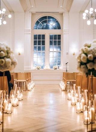 A brightly lit event space with white walls, a large window, and a wooden floor, set up for a ceremony with chairs, candles, and flowers.
