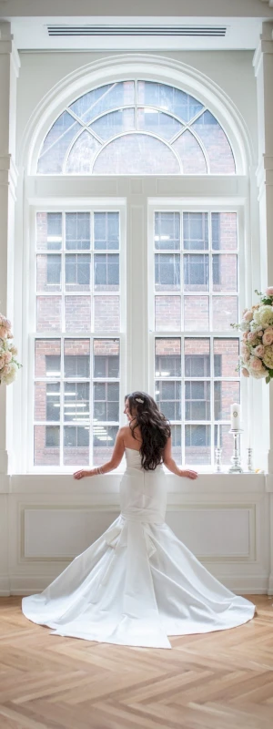 A bride in a white wedding dress standing in front of a large window with flowers on either side.