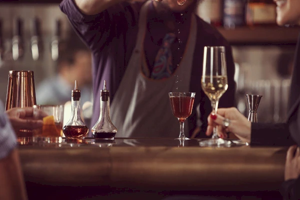 A bartender preparing a drink at a bar with two glasses and bottles of liquid on the counter.