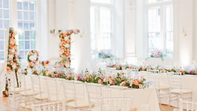 A brightly lit room with long tables set for dining, adorned with floral arrangements and white chairs.