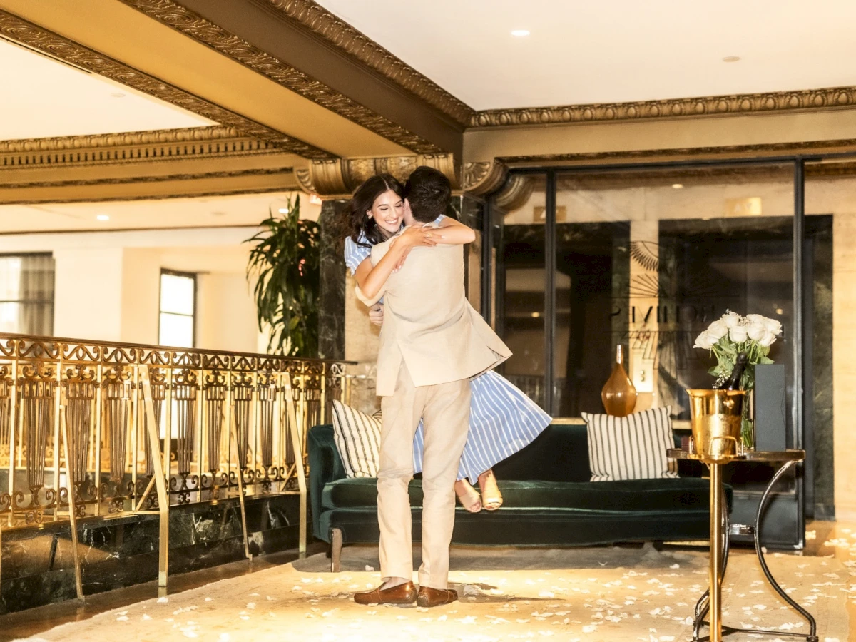 A couple embracing in a luxurious lobby with a green sofa, gold accents, and a balcony.