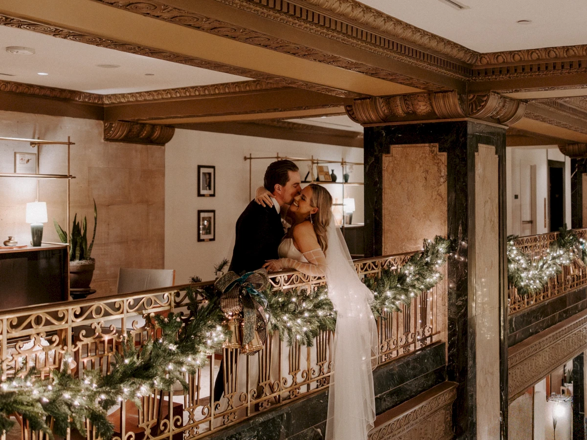 A couple in wedding attire kissing on a balcony with a garland and lights.