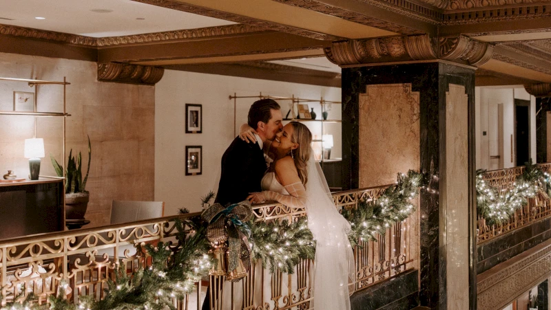 A couple in wedding attire embracing on a balcony with a gold railing and garland.