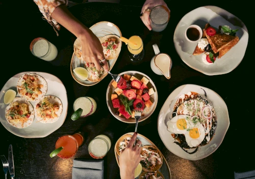 A table with various plates of food and drinks, with two people's hands reaching for food.