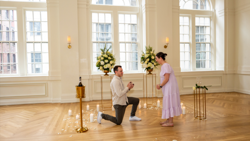 A man proposing to a woman in a beautifully decorated lobby with large windows, floral arrangements, and candles.