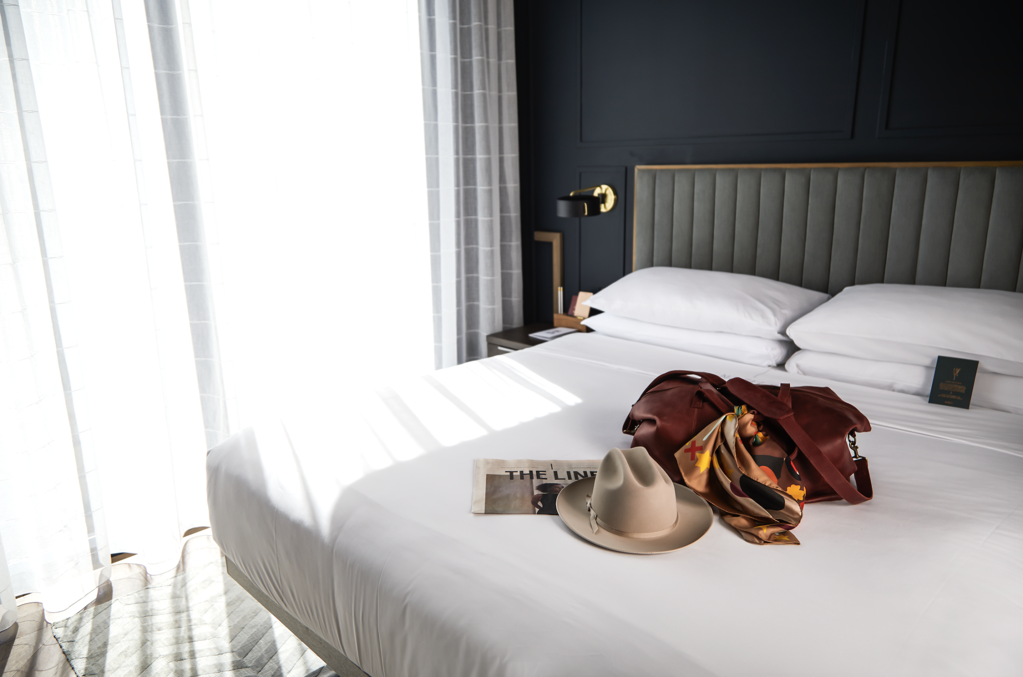 A neatly made bed with a hat, scarf, book, and pouch on top, next to a window with sunlight streaming in.