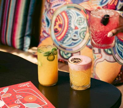 three colorful drinks on a table with a menu and a hand holding a drink in front of a colorful couch