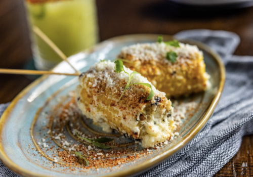 Grilled corn on the cob topped with cheese and spices on a plate, served on a textured cloth, with a drink in the background.