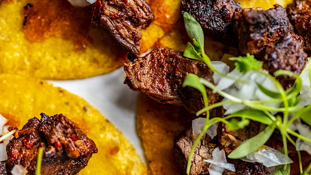 Tacos topped with grilled meat, diced onions, cilantro, and a lime wedge on the side are shown.
