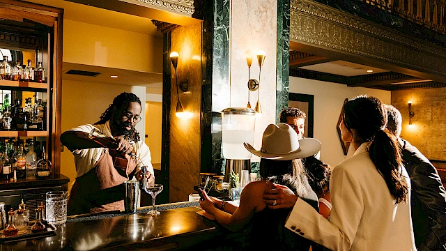 A bartender prepares drinks for a group in a warmly-lit, stylish bar setting.