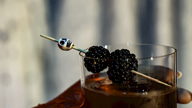 A person is holding a glass of dark-colored drink garnished with blackberries on a skewer, with partial henna design visible on the hand.