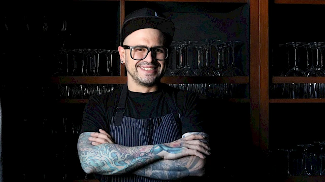 A person with tattoos wearing glasses and a cap, standing with arms crossed, in front of shelves with glassware, smiling.