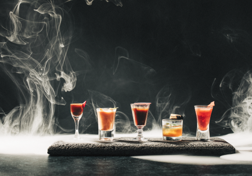 A selection of five cocktails in diverse glassware is artfully arranged on a smoky background, creating a mysterious and elegant scene.