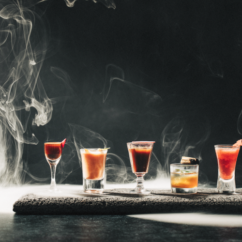 A selection of five cocktails in diverse glassware is artfully arranged on a smoky background, creating a mysterious and elegant scene.