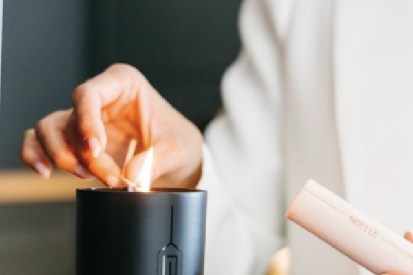 A person in a white outfit lights a candle in a black holder using a slim pink lighter on a wooden surface.