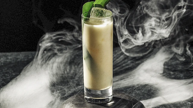 A tall glass filled with a creamy drink, garnished with green leaves, surrounded by swirling smoke on a dark surface.