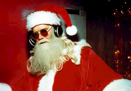 Santa Claus is wearing sunglasses and headphones, standing in front of a festive background with Christmas lights and decorations.