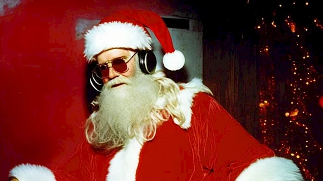 Santa Claus is wearing sunglasses and headphones, standing in front of a festive background with Christmas lights and decorations.