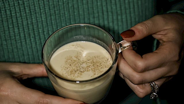 A person wearing a green sweater and gold necklace holds a transparent cup of frothy coffee with red painted nails.