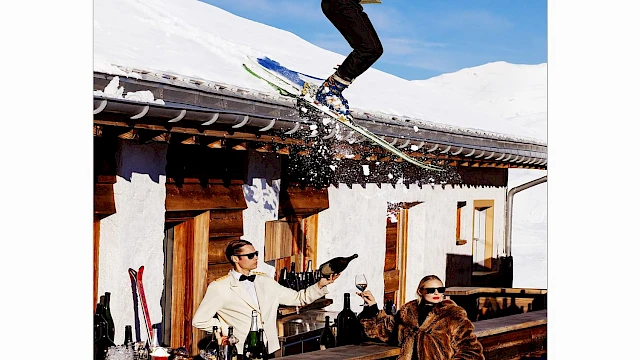 A skier jumps off a snowy roof holding a drink tray, while two people below enjoy drinks at an outdoor bar in a winter setting.