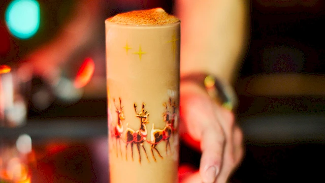A festive cocktail in a decorated glass on a bar counter with colorful lights in the background and a person reaching towards it.