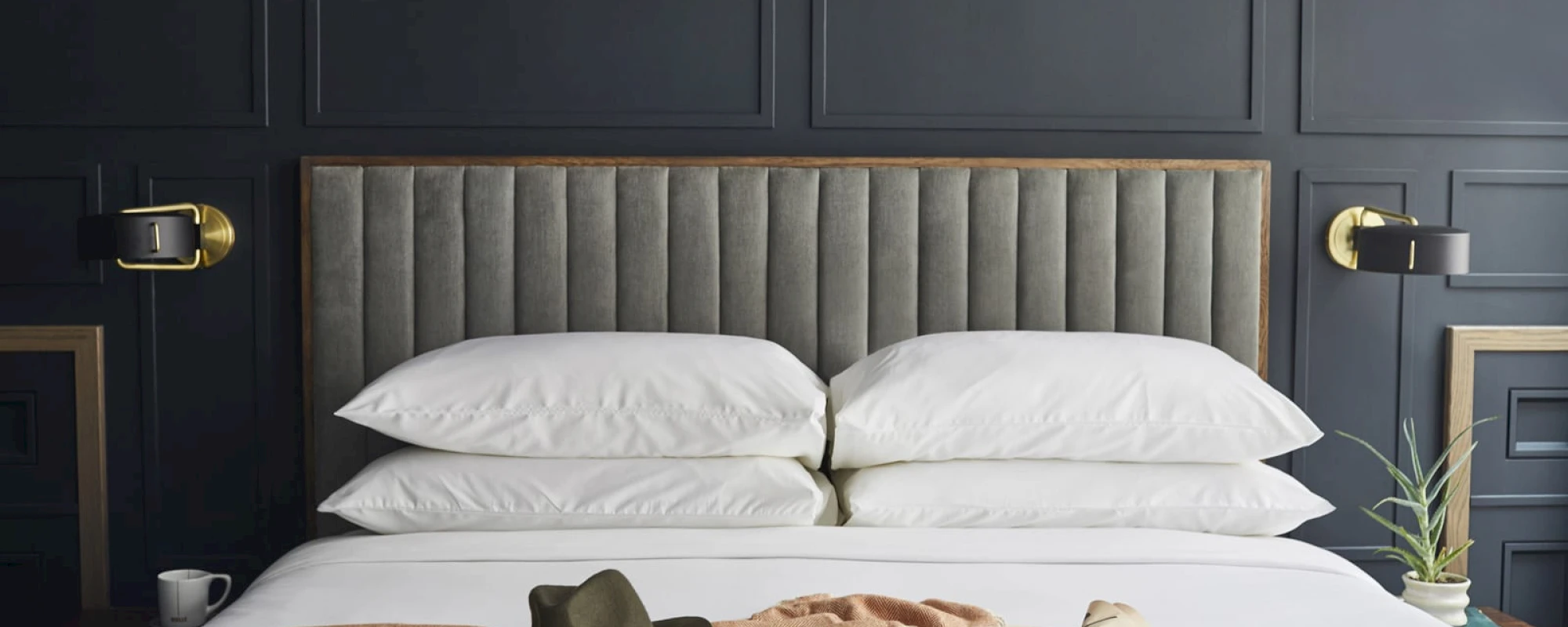 A bed with white bedding and a gray headboard, flanked by gold lights and a plant and mug on either side.