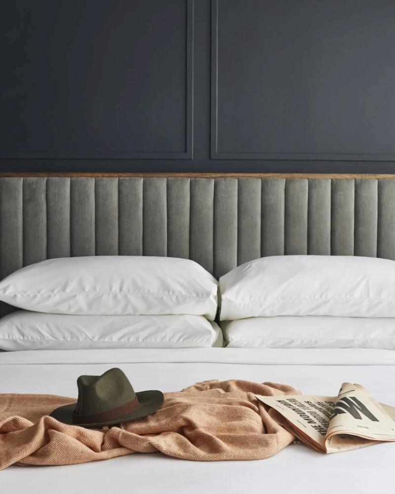A close-up of a bed with a gray headboard, white pillows, and a brown blanket with a hat and book on it.