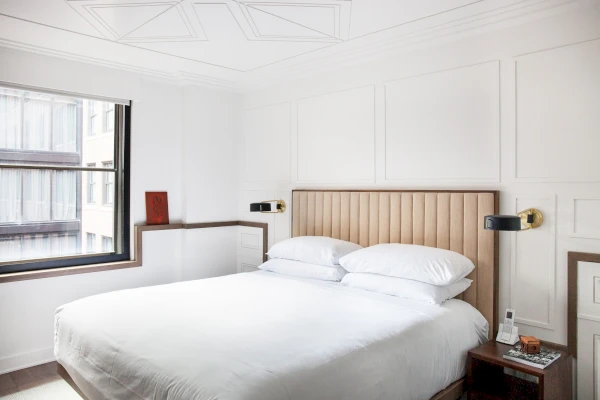 A white hotel room with a large bed, wooden headboard, and a window