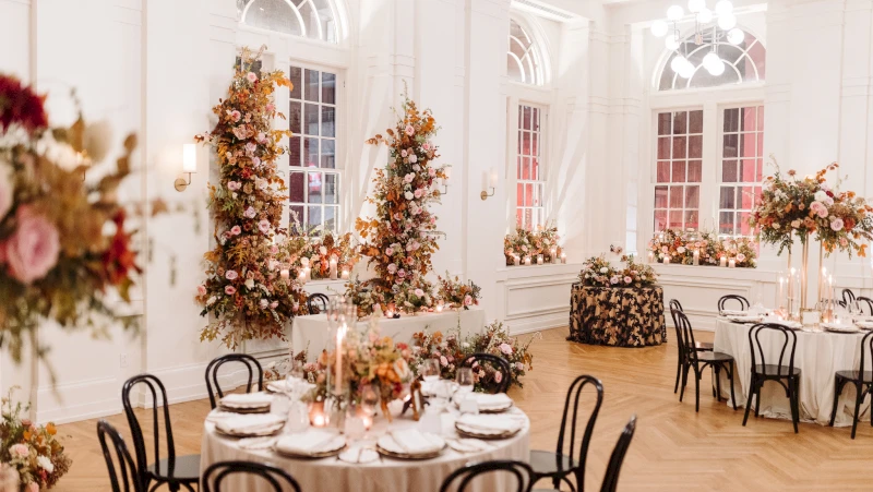A decorated dining room with round tables, white tablecloths, black chairs, floral arrangements, and candles.