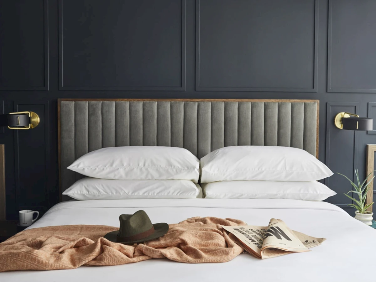 A neatly made bed with white bedding, a gray upholstered headboard, and gold sconce lights on either side.