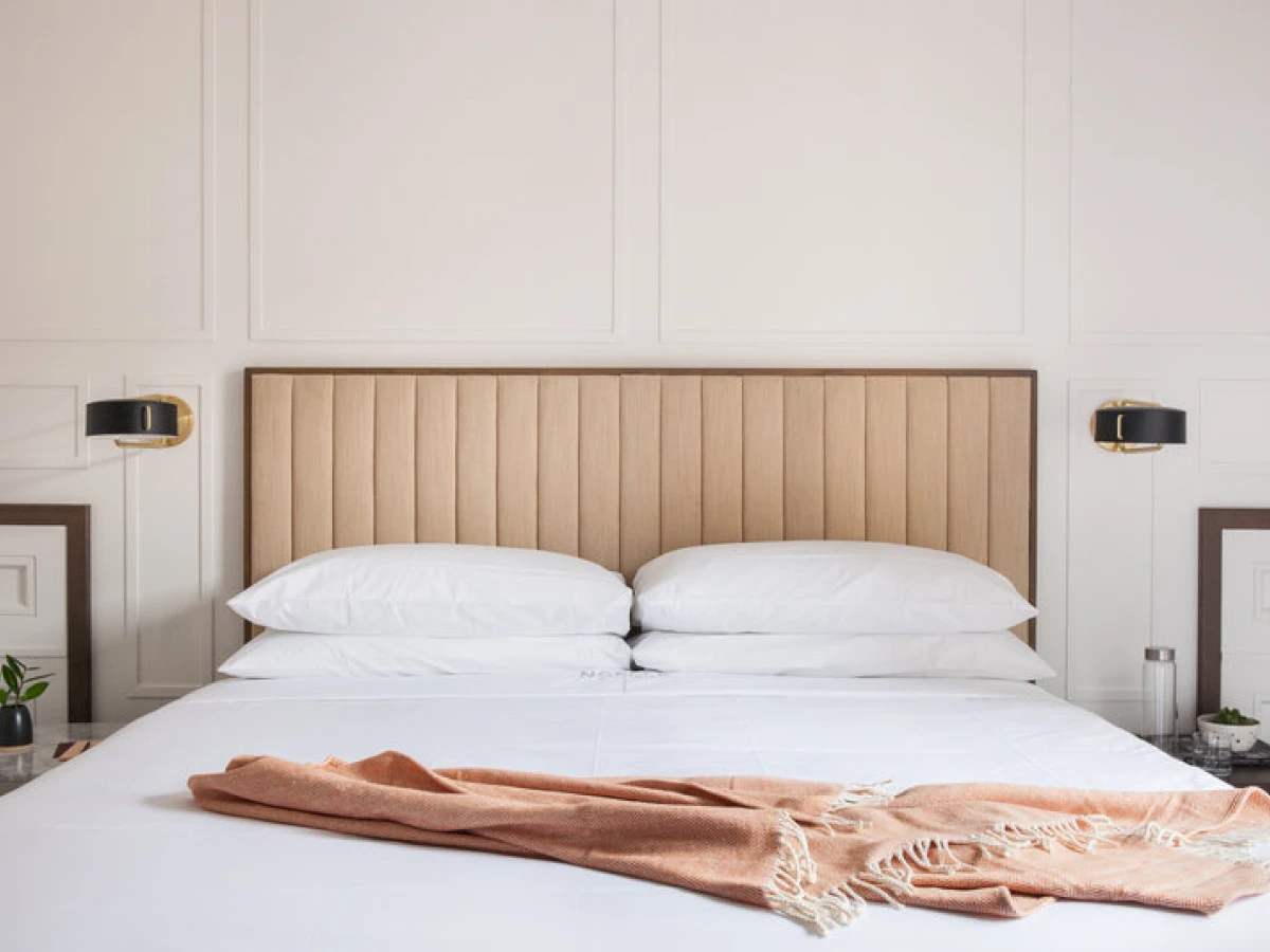 A neatly made bed with white bedding, two pillows, and a tan throw blanket at the foot, against a light wood headboard with vertical paneling, flanked by black wall lights and nightstands.