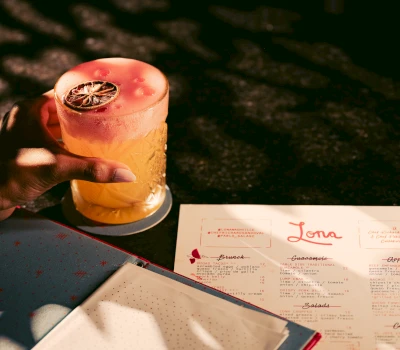 A hand holding a cocktail with a pink foam top and a slice of lime, on a table with a menu.