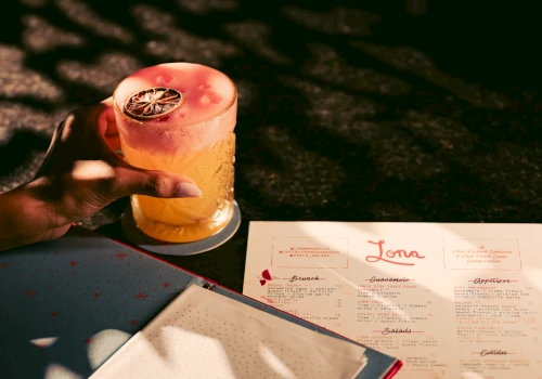 A hand holding a cocktail with a pink top and a menu on the table.
